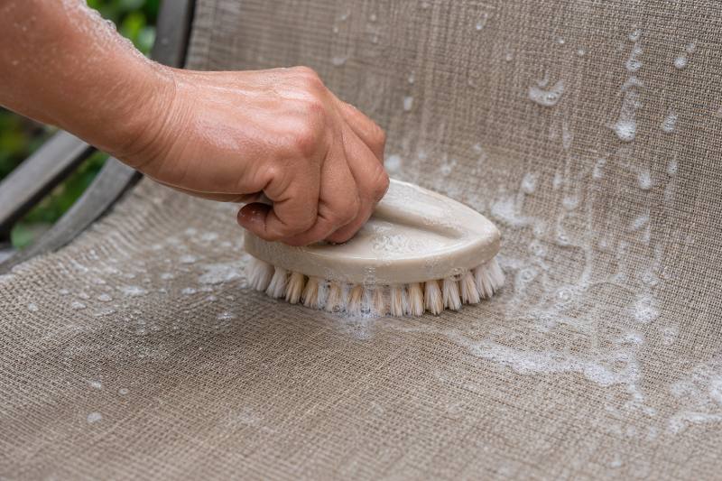Loveseat Sling Care: 7 Proven Tips for Extended Outdoor Life 4 Close-up of hands gently cleaning a loveseat sling with a soft brush and cleaning solution, emphasizing proper Loveseat Sling Care techniques. Outdoor setting.